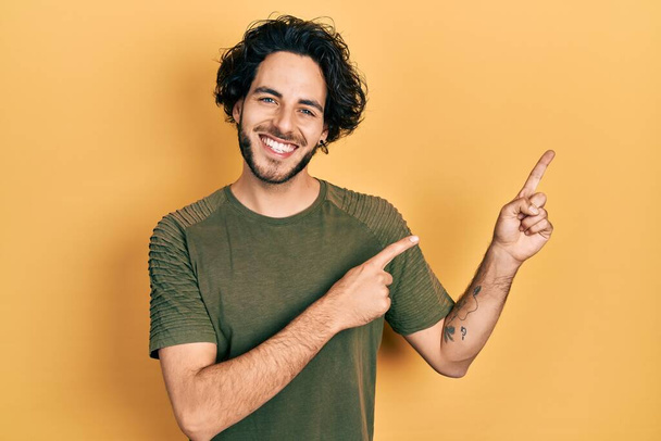 Schöner hispanischer Mann in lässigem grünem T-Shirt, der lächelnd in die Kamera blickt und mit zwei Händen und Fingern zur Seite zeigt.  - Foto, Bild