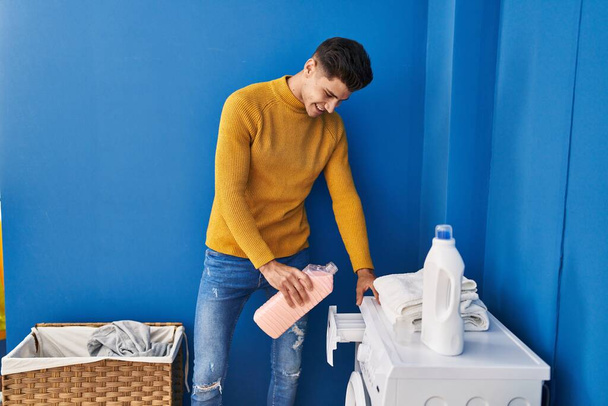 Jovem hispânico sorrindo confiante derramando detergente na máquina de lavar roupa na lavanderia - Foto, Imagem