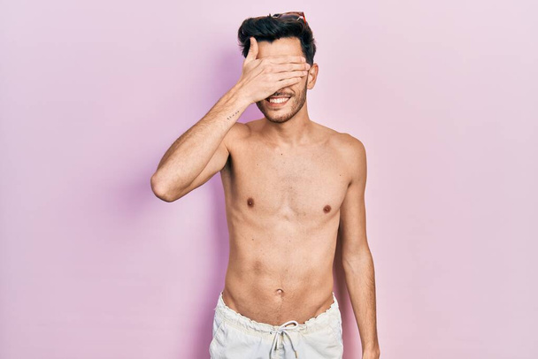 Junger hispanischer Mann in Badebekleidung ohne Hemd, lächelnd und lachend mit der Hand auf dem Gesicht, die zur Überraschung die Augen verdeckt. Blindes Konzept.  - Foto, Bild