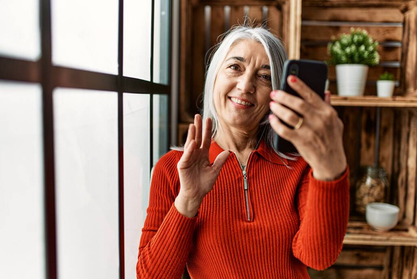Keski-ikä harmaa tukka nainen hymyilee luottavainen ottaa videopuhelu kotona - Valokuva, kuva