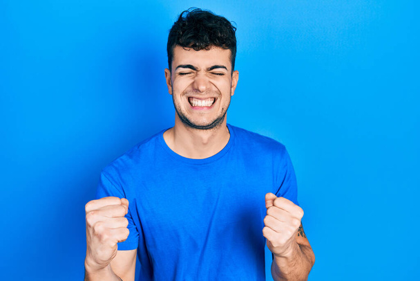 Junger hispanischer Mann in lässigem blauem T-Shirt mit erhobenen Armen und geschlossenen Augen, der den Sieg lächelnd feiert. Siegerkonzept.  - Foto, Bild
