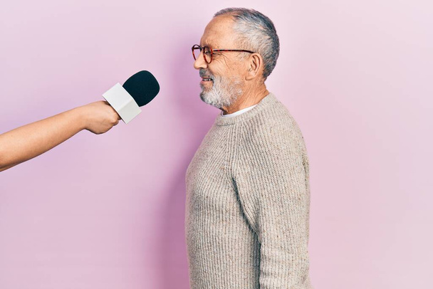 Красивый пожилой человек с бородой интервью с репортером держа микрофон глядя в сторону, расслабиться профиль позировать с естественным лицом с уверенной улыбкой.  - Фото, изображение