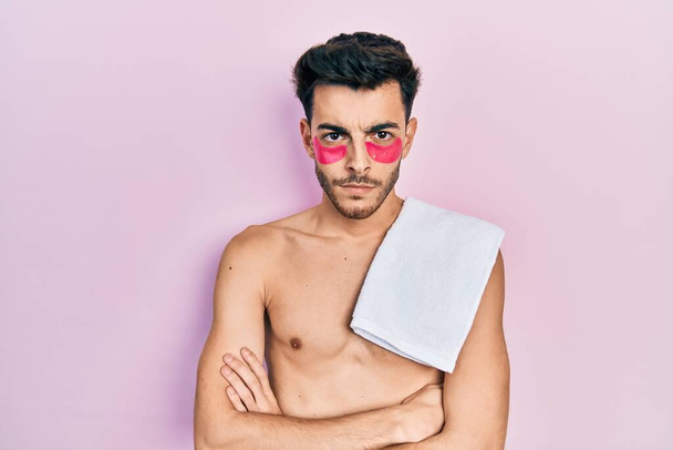 Jonge Spaanse man shirtloos het dragen van handdoek en oogzakken patches sceptisch en nerveus, afkeurende uitdrukking op gezicht met gekruiste armen. negatieve persoon.  - Foto, afbeelding