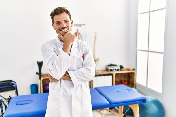 Handsome young man working at pain recovery clinic looking confident at the camera smiling with crossed arms and hand raised on chin. thinking positive.  - Foto, imagen