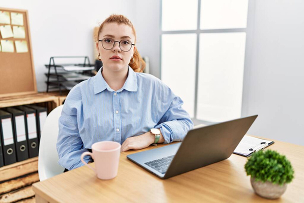 Jeune rousse femme travaillant au bureau à l'aide d'un ordinateur portable détendu avec une expression sérieuse sur le visage. simple et naturel regardant la caméra.  - Photo, image