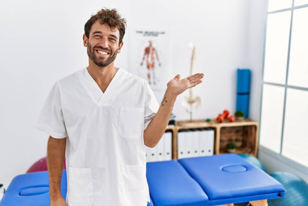 Jonge knappe fysiotherapeut werkt bij pijn herstel kliniek glimlachende vrolijke presentatie en wijzen met palm van de hand te kijken naar de camera.  - Foto, afbeelding