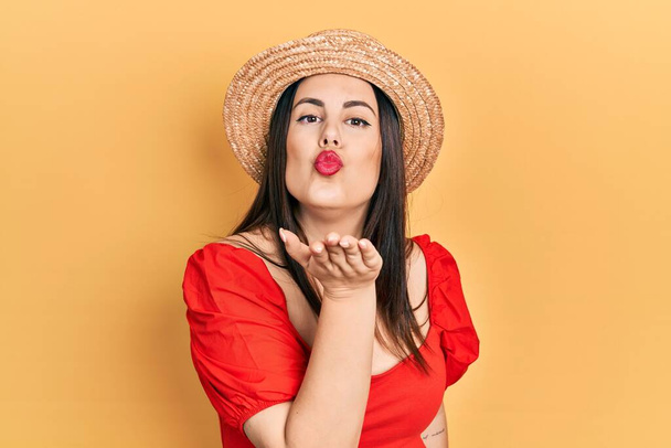 Mujer hispana joven con sombrero de verano mirando a la cámara soplando un beso con la mano en el aire siendo encantadora y sexy. expresión de amor.  - Foto, imagen