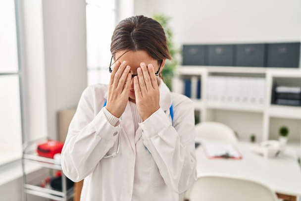 Egy fiatal barna doktornő sztetoszkópot visel a klinikán szomorú arckifejezéssel, miközben kezekkel takarja el az arcát, miközben sír. depresszió fogalma.  - Fotó, kép