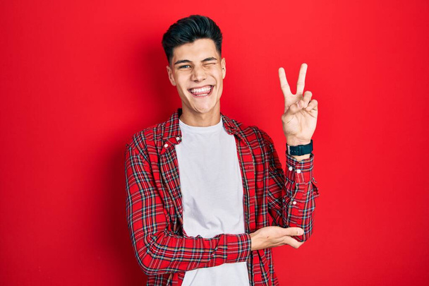 Jeune homme hispanique portant des vêtements décontractés souriant avec un sourire heureux clin d'oeil à la caméra faisant signe de victoire. numéro deux.  - Photo, image