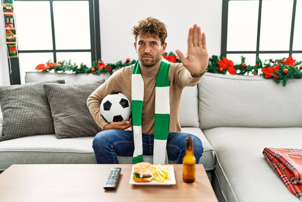 Νέοι ισπανόφωνοι χούλιγκαν ποδοσφαίρου κρατώντας μπάλα στο σπίτι με ανοιχτό χέρι κάνει στοπ υπογράψει με σοβαρή και σίγουρη έκφραση, χειρονομία άμυνας  - Φωτογραφία, εικόνα