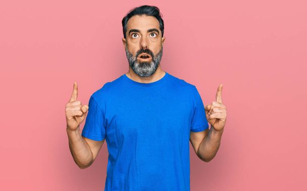 Mann mittleren Alters mit Bart und lässigem blauem T-Shirt staunt und überrascht und zeigt mit erhobenen Armen und Fingern nach oben.  - Foto, Bild