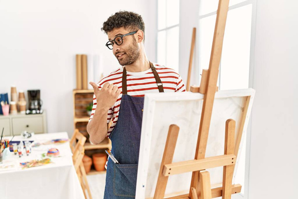Арабський молодий чоловік у художній студії вказує на спину з рукою і пальцями вгору, посміхаючись впевнено  - Фото, зображення