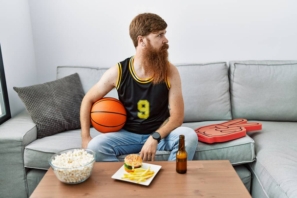 Homme caucasien à longue barbe tenant ballon de basket-ball acclamant jeu de télévision regardant vers le côté, pose de profil relax avec le visage naturel et sourire confiant.  - Photo, image