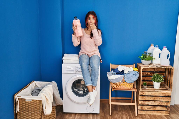 Junge kaukasische Frau sitzt auf der Waschmaschine und hält eine Spülmittelflasche in der Hand, die den Mund mit der Hand bedeckt, schockiert und ängstlich vor Irrtum. Überraschte Miene  - Foto, Bild