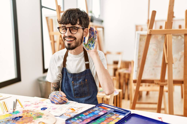 Homme hispanique avec barbe au studio d'art souriant avec la main sur l'oreille en écoutant une rumeur ou des commérages. concept de surdité.  - Photo, image