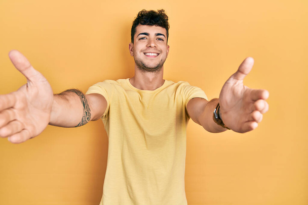 Junger hispanischer Mann in lässigem T-Shirt, der mit offenen Armen in die Kamera lächelt und sich umarmt. fröhlicher Ausdruck, der das Glück umarmt.  - Foto, Bild
