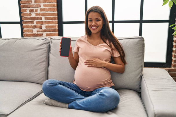 Elinde akıllı telefon tutan genç hamile bir kadın pozitif ve mutlu görünüyor. Dişlerini gösteren bir gülümsemeyle ayakta duruyor ve gülümsüyor.  - Fotoğraf, Görsel