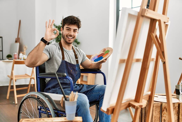 Young hispanic man sitting on wheelchair painting at art studio smiling positive doing ok sign with hand and fingers. successful expression.  - Zdjęcie, obraz