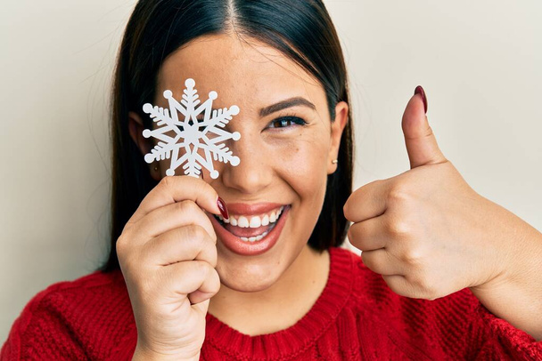 Beautiful brunette woman holding snowflake over eye smiling happy and positive, thumb up doing excellent and approval sign  - Photo, Image