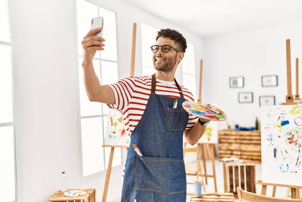 Молодой араб-художник улыбающийся счастливый делает селфи на смартфоне в арт-студии. - Фото, изображение