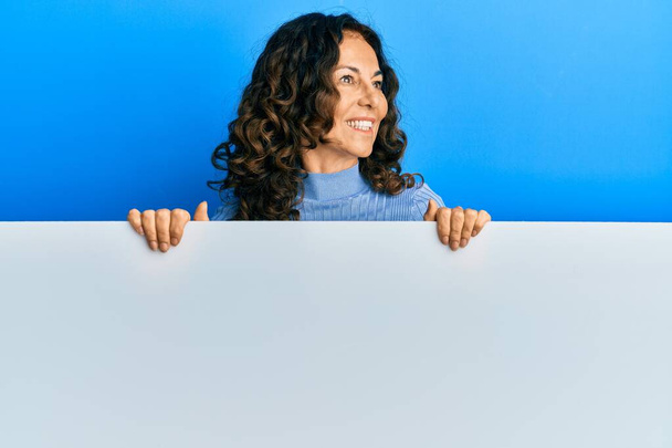 Eine hispanische Frau mittleren Alters hält ein leeres Transparent in der Hand, lächelt zur Seite und starrt nachdenklich weg.  - Foto, Bild