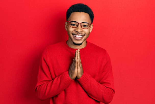Fiatal afro-amerikai férfi alkalmi ruhát és szemüveget visel, kéz a kézben imádkozik, bocsánatért könyörög, magabiztosan mosolyog..  - Fotó, kép