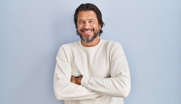 Knappe man van middelbare leeftijd met een casual trui over een blauwe achtergrond vrolijk gezicht glimlachend met gekruiste armen kijkend naar de camera. positieve persoon.  - Foto, afbeelding