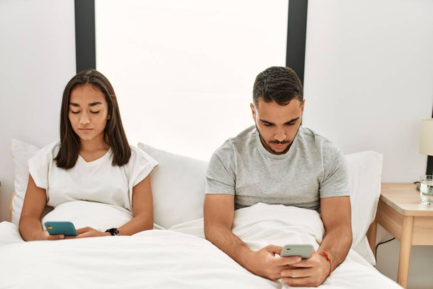 Joven pareja latina usando smartphone acostado en la cama en casa. - Foto, imagen