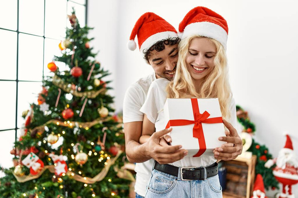 Fiatal pár visel karácsonyi kalap ölelés és gazdaság ajándék otthon. - Fotó, kép