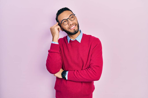 Hispanischer Mann mit Bart, Businesshemd und Brille mit der Hand am Kinn, der über Fragen nachdenkt, nachdenklichen Ausdruck. lächelndes und nachdenkliches Gesicht. Zweifelhaftes Konzept.  - Foto, Bild