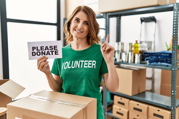 Middle age woman wearing volunteer t shirt holding please donate banner surprised with an idea or question pointing finger with happy face, number one  - Foto, Imagen