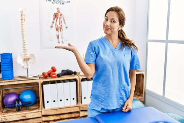 Middelbare leeftijd Spaanse fysiotherapeut vrouw werken bij pijn recovery kliniek glimlachende vrolijke presentatie en wijzen met palm van de hand te kijken naar de camera.  - Foto, afbeelding