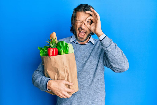 Красивый мужчина средних лет держит бумажный пакет с продуктами, улыбаясь счастливо делает хорошо знак с рукой на глазу, глядя сквозь пальцы  - Фото, изображение