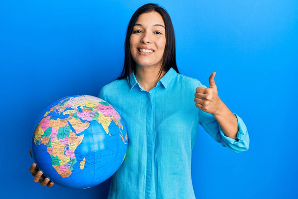 Junge lateinische Frau hält großen Weltball lächelnd glücklich und positiv, Daumen hoch dabei hervorragende und Zustimmung Zeichen  - Foto, Bild