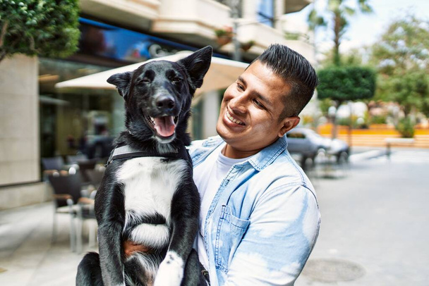 Молодий латинський чоловік посміхається щасливий тримає собаку в місті
. - Фото, зображення