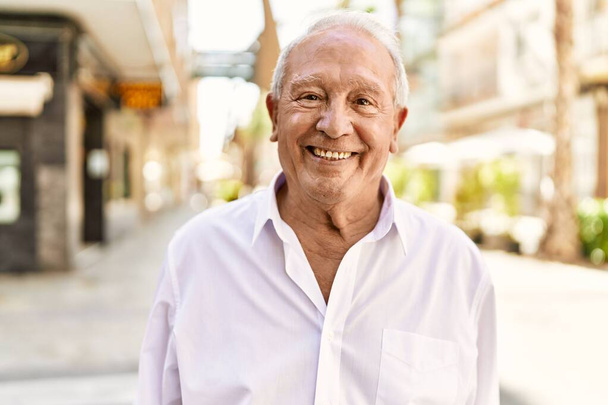 Hombre mayor con el pelo gris de pie feliz al aire libre en un día soleado. Sonriendo feliz a los 70 para la jubilación. - Foto, imagen