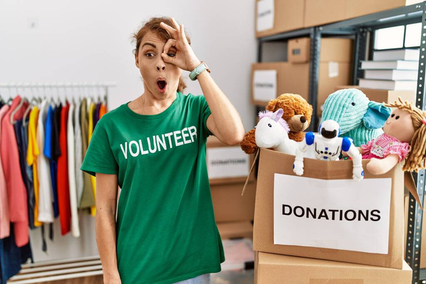 Hermosa mujer caucásica vistiendo una camiseta voluntaria en el stand de donaciones haciendo un gesto bien sorprendido con la cara sorprendida, los ojos mirando a través de los dedos. expresión incrédula.  - Foto, imagen