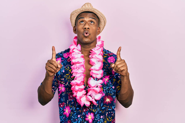 Young black man wearing summer shirt and hawaiian lei amazed and surprised looking up and pointing with fingers and raised arms.  - Photo, Image