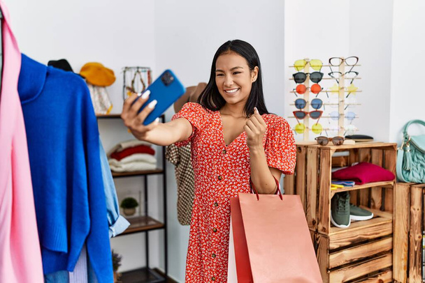 Jeune femme latine faire selfie par le smartphone tenant des vêtements au magasin de vêtements - Photo, image