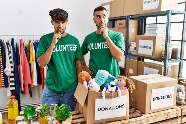 Νεαρό ομοφυλόφιλο ζευγάρι που φοράει εθελοντικό μπλουζάκι σε δωρεές στέκεται σκεπτόμενο ανησυχώντας για μια ερώτηση, ανήσυχος και νευρικός με το χέρι στο πηγούνι  - Φωτογραφία, εικόνα