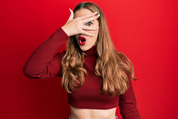 Young blonde woman wearing turtleneck sweater peeking in shock covering face and eyes with hand, looking through fingers with embarrassed expression.  - Fotografie, Obrázek