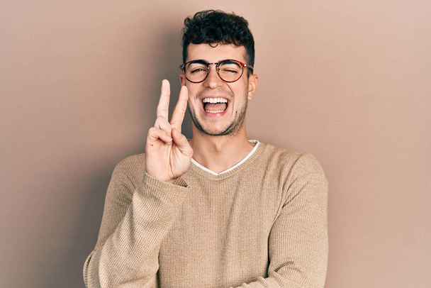 Νεαρός Ισπανός που φοράει καθημερινά ρούχα και γυαλιά χαμογελώντας με χαρούμενο πρόσωπο να κλείνει το μάτι στην κάμερα κάνοντας το σήμα της νίκης με τα δάχτυλα. Νούμερο δύο..  - Φωτογραφία, εικόνα