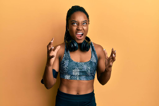 African american woman with braided hair wearing sportswear and arm band crazy and mad shouting and yelling with aggressive expression and arms raised. frustration concept.  - Fotoğraf, Görsel