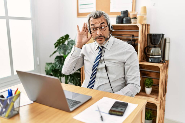 Hombre de negocios hispano de mediana edad que trabaja en la oficina con auriculares de operador sonriendo con la mano sobre el oído escuchando un rumor o chismes. concepto de sordera.  - Foto, imagen