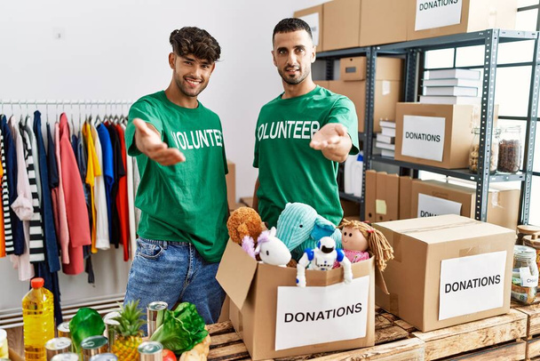 Young gay couple wearing volunteer t shirt at donations stand smiling cheerful offering palm hand giving assistance and acceptance.  - Photo, Image