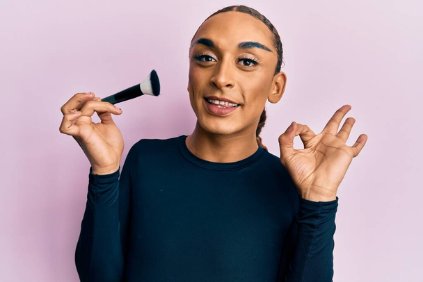 Hispanic man wearing make up and long hair holding makeup brush doing ok sign with fingers, smiling friendly gesturing excellent symbol  - Photo, Image