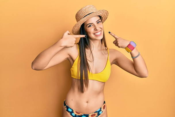 Junge hispanische Frau mit Bikini und Sommermütze lächelt fröhlich und zeigt mit den Fingern Zähne und Mund. Zahngesundheitliches Konzept.  - Foto, Bild