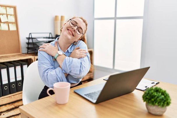 Young redhead woman working at the office using computer laptop hugging oneself happy and positive, smiling confident. self love and self care  - Foto, afbeelding