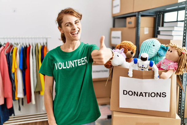 Beautiful caucasian woman wearing volunteer t shirt at donations stand approving doing positive gesture with hand, thumbs up smiling and happy for success. winner gesture.  - Photo, Image
