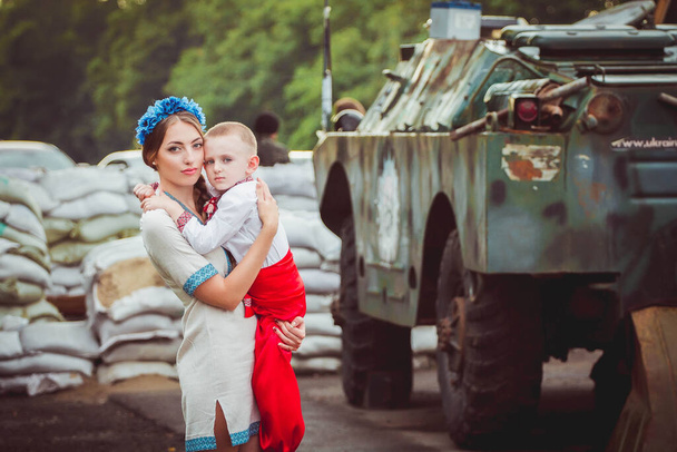 若いウクライナ人女性は、装甲兵員輸送車やサンドバッグを背景に、伝統的な民族衣装を着た子供の男の子と一緒に道端に立っている。ウクライナにおけるロシアの軍事侵攻の概念。ウクライナとヨーロッパでの戦争。子供と  - 写真・画像